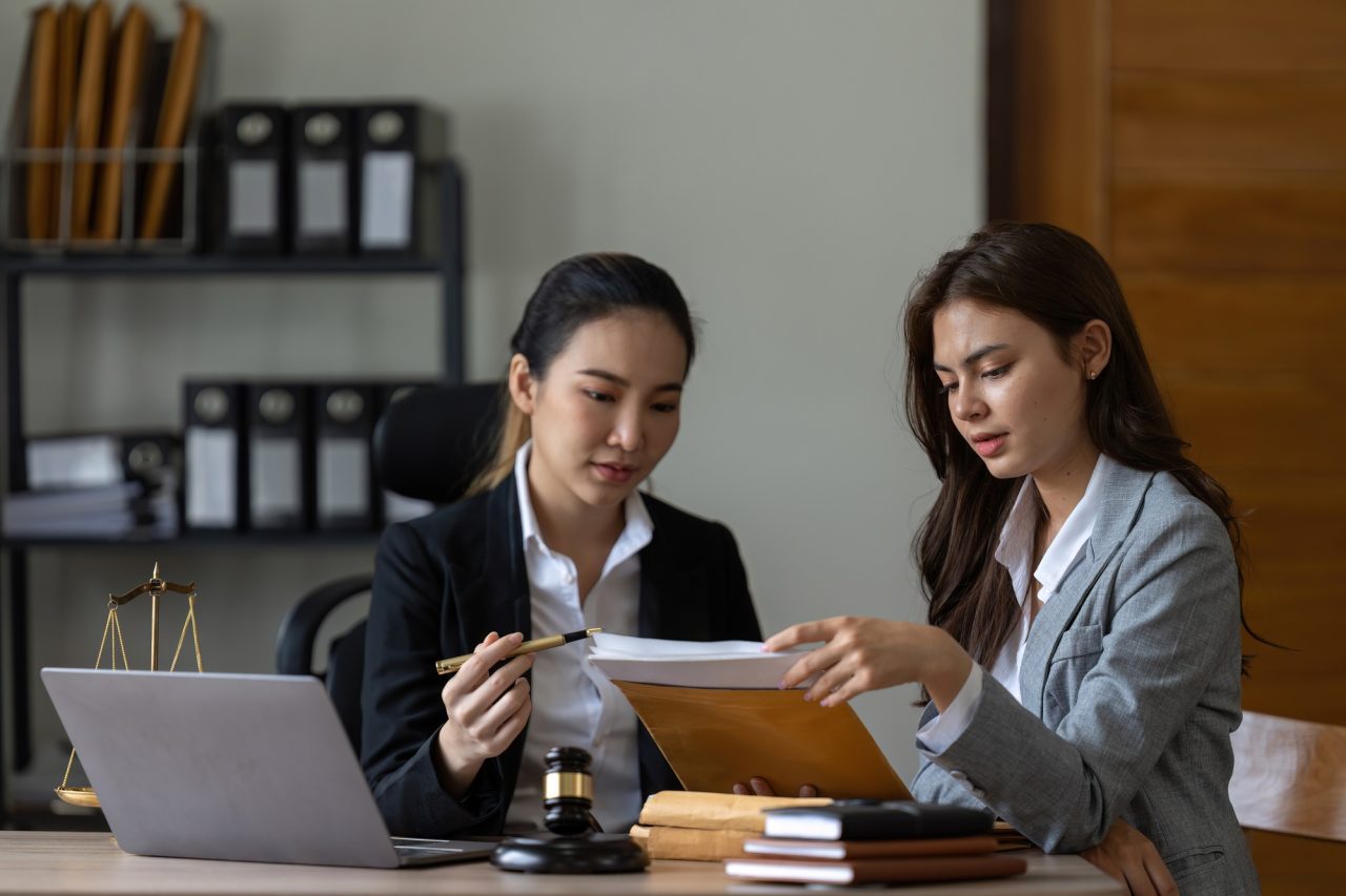 Two women are seated at a desk, discussing documents related to a legal matter. A laptop and a gavel are visible on the table, indicating a professional setting.