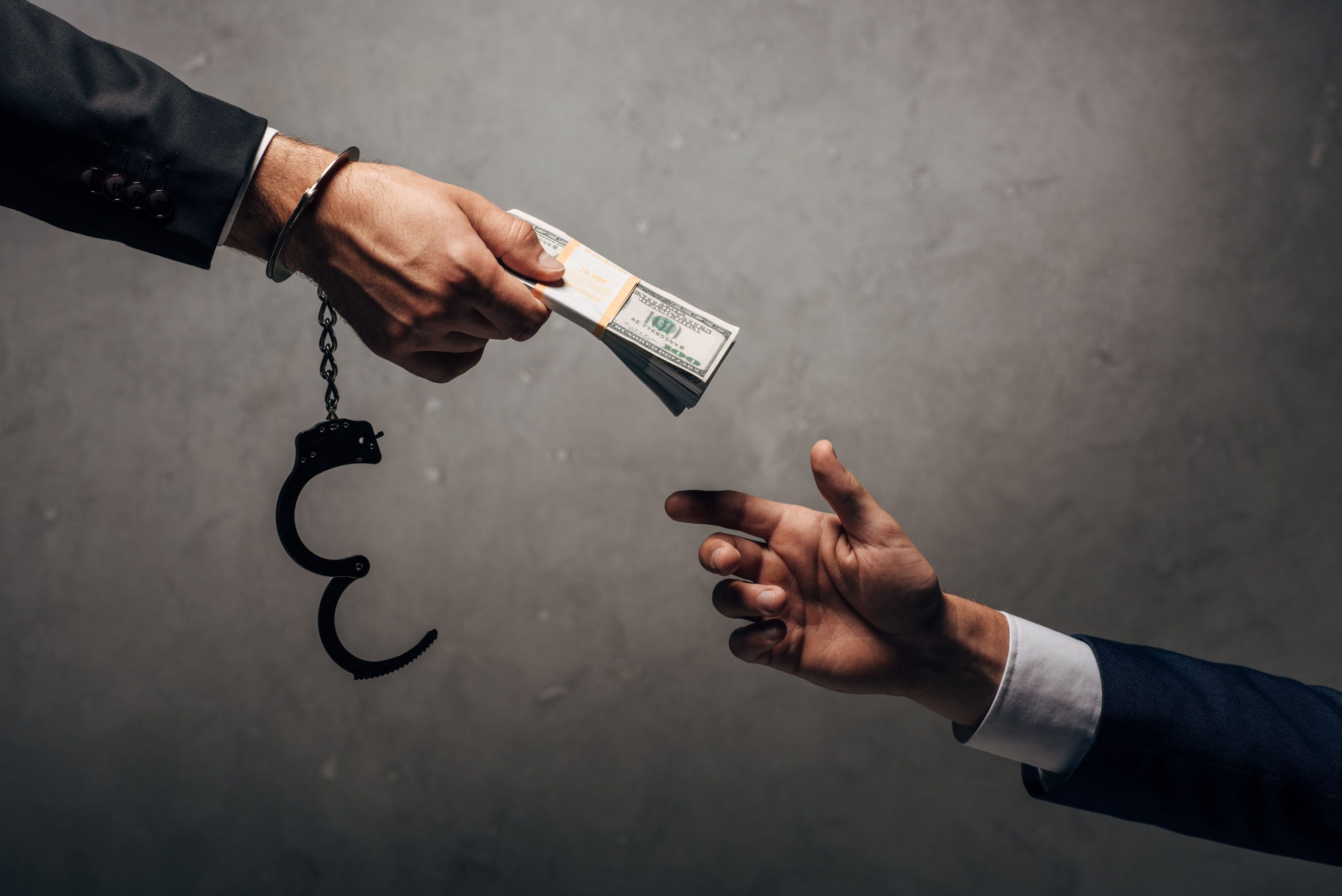 A hand in a suit reaches out to receive cash from another hand, which is attached to handcuffs. The background is a muted gray, emphasizing the transaction related to bail bonds.