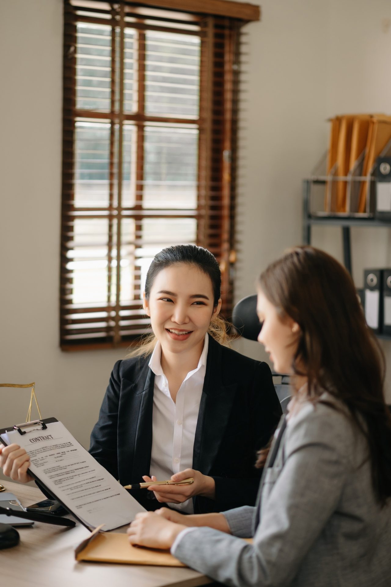 Two women are seated at a desk in an office, discussing documents. One woman is smiling and holding a clipboard, while the other listens attentively. Natural light filters through a window in the background.