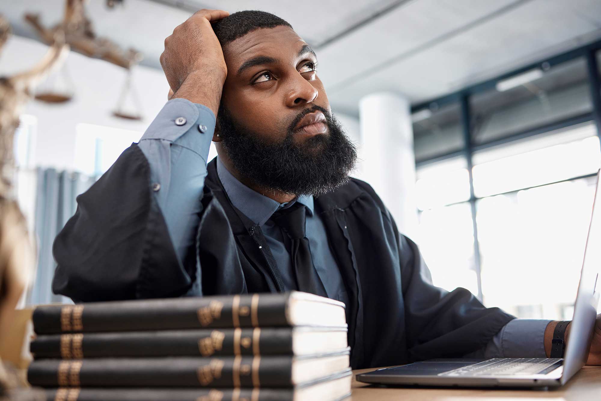 Bearded lawyer in a black robe sits thoughtfully at a desk beside stacked law books and an open laptop.